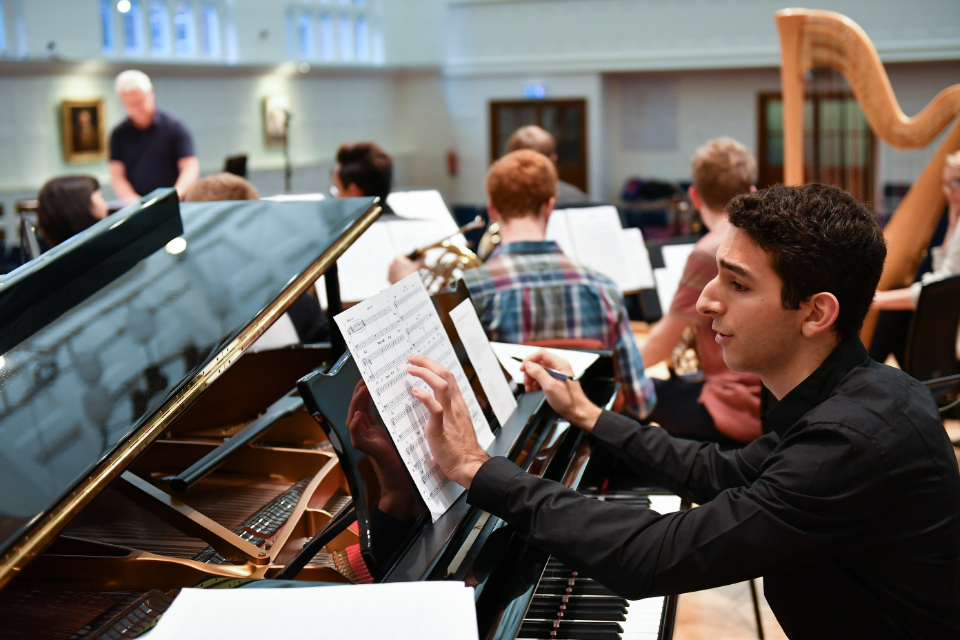 A male pianist, wearing a black shirt, making a note on his notepad next to his music sheet, with an orchestra rehearsal in the background, in a well-lit room.